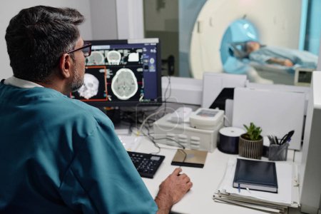 Selective focus over-the-shoulder shot of male radiographer controlling CT scanner using desktop computer