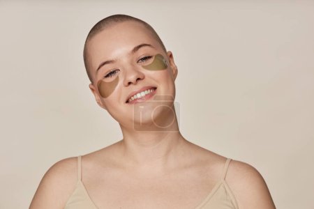 Studio portrait of happy young Caucasian woman with shaved head posing for camera with collagen eye patches on face