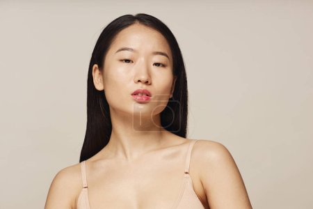 Medium closeup studio portrait of young Asian woman with long hair wearing beige bra looking at camera