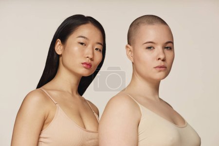 Studio portrait of self-confident young Asian and Caucasian women wearing neutral toned bras posing for camera