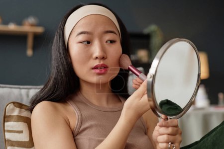 Young Asian woman holding mirror applying bronzer on face, medium closeup shot