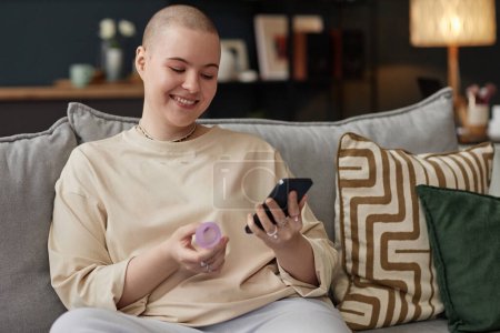 Medium portrait of happy young Caucasian woman with shaved head holding menstrual cup checking period calendar on smartphone