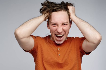 Medium closeup studio portrait of angry young Caucasian man wearing orange t-shirt pulling his hair out