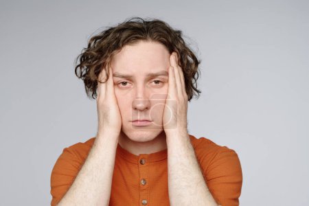 Medium closeup studio portrait of young tired man with curly hair posing for camera with hands covering his ears
