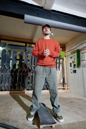 Vertical low angle long shot of young Caucasian man standing in skatepark posing for camera