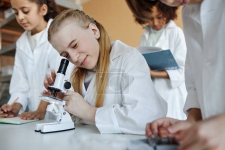 Selective focus shot of Caucasian schoolgirl sitting at desk in classroom using microscope during Chemistry lesson