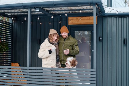 Caucasian man and woman in love wearing casual warm clothes standing on porch with their dog on cold winter day, copy space