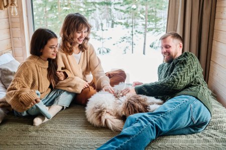 Caucasian man, woman and teen girl spending winter day indoors relaxing on bed with their dog in country house