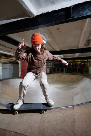Modern gen Z girl in streetwear training skateboarding in skatepark, vertical long shot