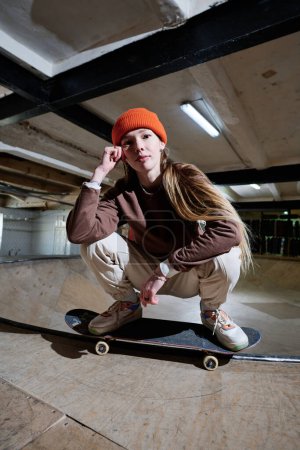 Vertical shot of young gen Z woman with long hair posing for camera squatting on skateboard, copy space