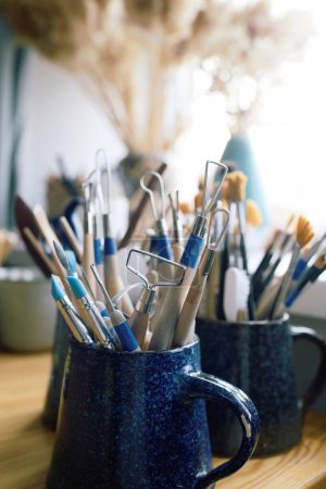 Gros plan de divers outils de sculpture en argile et pinceaux dans des tasses en céramique bleue sur la table dans l'atelier, espace de copie