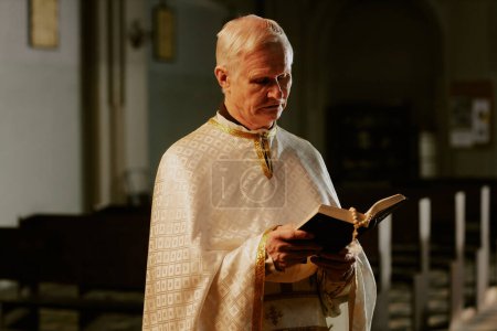 Medium shot of senior Caucasian Catholic priest standing indoors reading holy Bible book, copy space