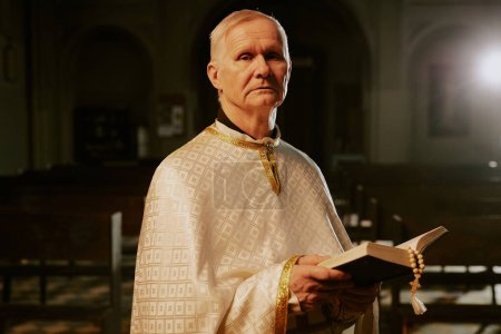 Medium portrait of elderly Catholic priest wearing beige chasuble standing indoors with Bible in hands looking at camera