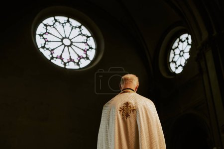 Rear view low angle shot of unrecognizable elderly Catholic priest wearing chasuble standing in church with round windows