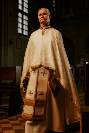 Vertical low angle full length shot of senior Catholic priest wearing liturgical clothes walking along church nave and looking at camera