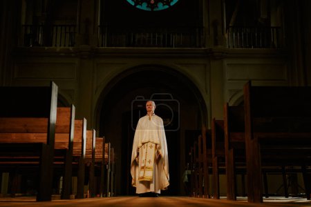 Wide shot of senior Caucasian Catholic priest wearing liturgical vestment standing in empty church, copy space