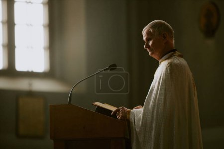 Senior Caucasian Catholic priest standing at lectern with mic opening Bible book, copy space