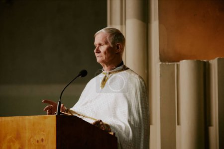Medium shot of elderly Caucasian Catholic priest standing at lectern speaking to parishioners during Church service