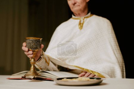 Selektive Fokusaufnahme eines hochrangigen kaukasischen katholischen Pastors, der am Abendmahlstisch steht und eine Tasse Wein in der Hand hält