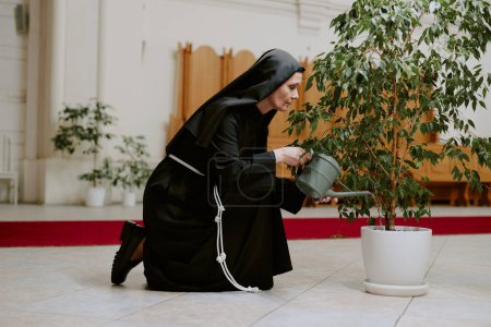 Seitenansicht einer älteren kaukasischen Nonne, die Ficus-Baum gießt, während sie sich um Pflanzen in der katholischen Kirche kümmert, Kopierraum