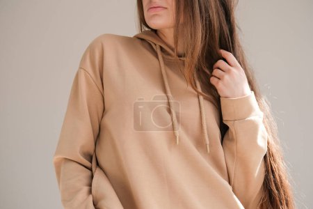 A female model posing in a beige hooded shirt touching long hair by a hand. A woman wearing a variety of basic clothing. Mock up. Concept ideas. Close up of clothing. Minimal style. Simplicity.