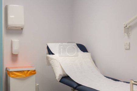 Eine erhöhte medizinische Couch im Innenraum, die das weiße Kopfkissen und die Papierbettrolle mit einfachen Wandspendern und Abfalleimern in den Fokus rückt.