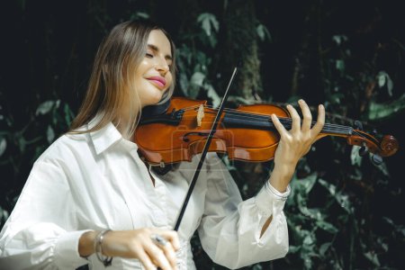 Photo for Close up portrait of smiling Caucasian woman playing violin in tropical forest. Closed eyes. Music and art concept. Female with blond hair wearing white dress. Background of lush green tropical leaves - Royalty Free Image