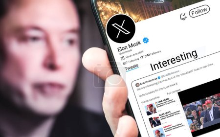 Foto de Poltava, Ucrania - 17 de marzo de 2024: Página de perfil de Elon Musk en Twitter con un smartphone en la mano. En el fondo hay un retrato desenfocado de Elon Musk. - Imagen libre de derechos