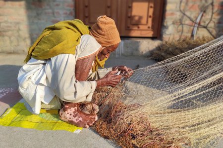 Foto de A very old but capable exemplary man of India making fishing nets in the sun on a winter morning wrapped in a blanket - Imagen libre de derechos