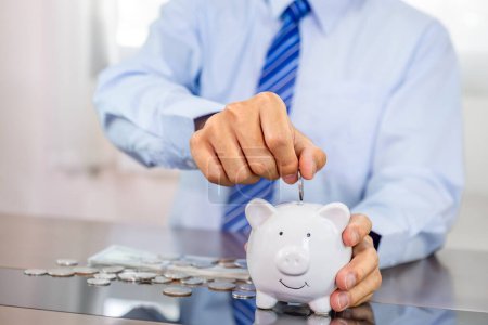 Businessman hand putting coin into blue piggy bank, saving money for future investment plan and retirement fund concept.