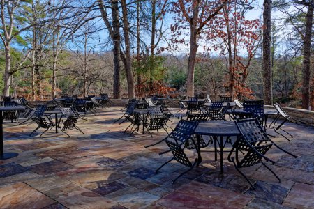 Téléchargez les photos : Un restaurant extérieur patio dans un parc fermé avec table en métal extérieur et chaises en grappes sur un sol plat en pierre coloré et avec des lumières suspendues et entouré par les arbres par une journée ensoleillée - en image libre de droit