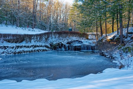 Téléchargez les photos : Une cascade coulante avec des glaçons se déversant dans un étang gelé entouré de neige fraîche à l'ombre avec le coucher du soleil en arrière-plan sur un terrain de golf en hiver - en image libre de droit