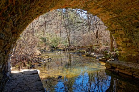 Standing on a ledge underneath inside the arch of an old stone bridge looking out at a tranquil creek in wintertime on a bright sunny day