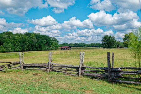 Téléchargez les photos : Une clôture rustique en bois construite à la main avec barbelés autour de la terre agricole avec une vieille grange en arrière-plan dans le pâturage par une journée ensoleillée avec des nuages blancs pelucheux à la fin de l'été - en image libre de droit