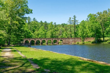 A landscape view of the walking path with stone benches and a fishing dock along the lake with the seven arch old stone bridge in the background on a bright sunny day in springtime