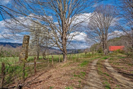 Photo for An old homestead only a chimney is left standing on farmland with a red roof barn on a dirt gravel backroad in the mountains of Tennessee in early springtime - Royalty Free Image