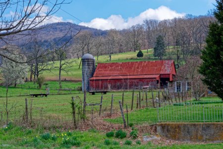 Alteingesessener Bauernhof mit leuchtend rot gestrichener Scheune mit Silo mit anderen Strukturen und Heuwagen, umgeben von rustikalem Zaun mit Bergen mit kahlen Bäumen im Hintergrund im Frühling