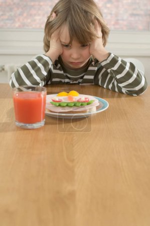 Frustrated boy sitting at table in front of healthy sandwich with vegetables and glass of tomato juice