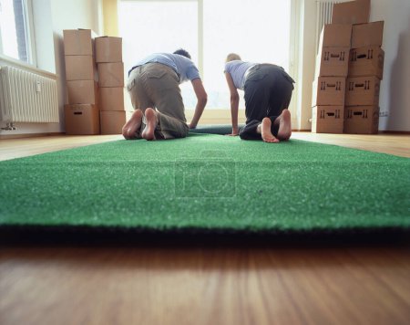 Photo for View taken from behind of couple kneeling on floor rolling out carpet in new home amidst stacks of cardboard boxes - Royalty Free Image