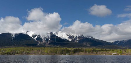 Photo for Snow-capped mountain ridge of Hudson Bay Mountain with white puffy clouds in a blue sky and the lake in the foreground; Smithers, British Columbia, Canada - Royalty Free Image