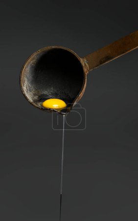Photo for Close-up of egg-white being separated from the egg yolk and dripping out of an old, rusted ladle on a dark background - Royalty Free Image