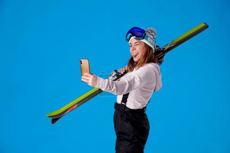 Mädchen mit Skiern und Schneebekleidung blickt auf ihr Handy und macht ein Selfie mit der Zunge vor blauem Hintergrund.