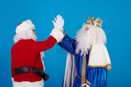 Photo for Santa Claus and a Wise Man high-fiving each other. Give my five on a blue background - Royalty Free Image