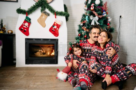 a family, with the father, mother, and daughter, wearing matching Christmas pajamas, taking family photos in front of the fireplace and a Christmas tree. The scene represents the joy, love, and togetherness of the holiday season