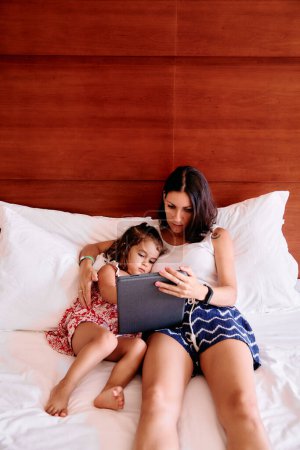 Photo for A little girl and her young mother have fun using an iPad on their hotel room bed - Royalty Free Image