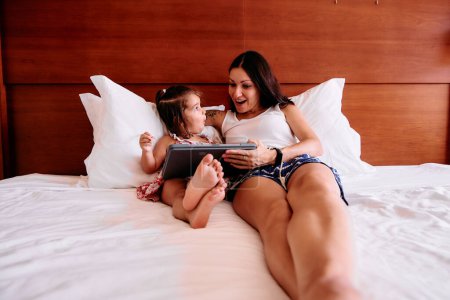 Photo for A little girl and her young mother have fun using an iPad on their hotel room bed - Royalty Free Image