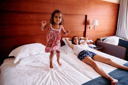 Photo for A little girl and her young mother have fun jumping on a hotel bed during their summer vacation. - Royalty Free Image