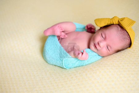 Foto de Primer plano de una niña recién nacida, dentro de una bola de lana azul sobre una manta amarilla con un lazo amarillo en la cabeza, durmiendo y riendo. - Imagen libre de derechos