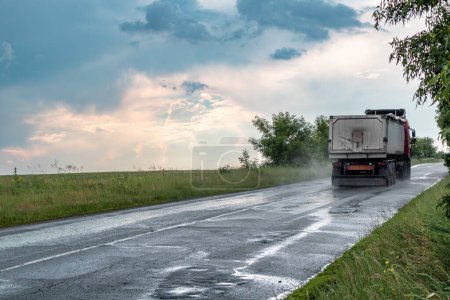 Téléchargez les photos : Véhicule lourd conduisant l'allée de route asphaltée humide par temps pluvieux orageux avec des nuages épiques et un paysage vert. Vue arrière de la voiture avec brouillard d'eau sur roues - en image libre de droit