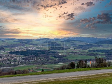 Stunning sunset over a lush Swiss landscape, featuring a winding path through vibrant green fields leading towards the majestic snow-capped Alps, under a dramatic sky painted with hues of orange and blue